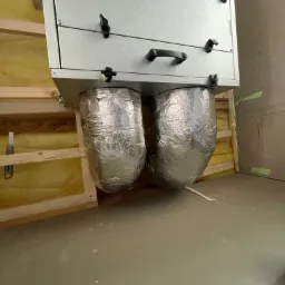 Szara centrala rekuperacyjna zamontowana pod sufitem z dwoma izolowanymi kanałami wentylacyjnymi, widoczne elementy konstrukcji drewnianej i izolacji oraz fragment podłogi.