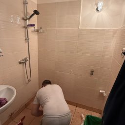 DOMINIX & ORIANIX & TRAIDING SPÓŁKA Z OGRANICZONĄ ODPOWIEDZIALNOŚCIĄ - Praca glazurnika w trakcie układania płytek w łazience w Warszawie. Widoczny fragment prysznica, umywalki i narzędzia pracy.