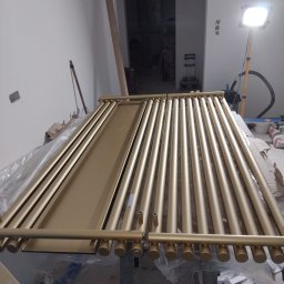 DOMINIX & ORIANIX & TRAIDING SPÓŁKA Z OGRANICZONĄ ODPOWIEDZIALNOŚCIĄ - Złoty kolektor słoneczny w trakcie montażu, widoczny w surowym wnętrzu z lampą budowlaną. Elementy instalacji ułożone na folii ochronnej, przygotowane do instalacji.