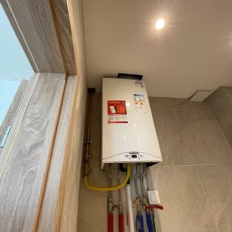 DOMINIX & ORIANIX & TRAIDING SPÓŁKA Z OGRANICZONĄ ODPOWIEDZIALNOŚCIĄ - Biały, wiszący piec gazowy Ariston w rogu łazienki z beżowymi płytkami. Widoczne rury instalacyjne w kolorach czerwonym i niebieskim. Oświetlenie sufitowe.