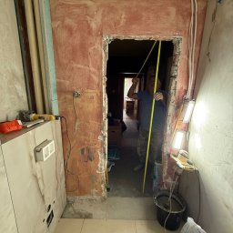 DOMINIX & ORIANIX & TRAIDING SPÓŁKA Z OGRANICZONĄ ODPOWIEDZIALNOŚCIĄ - Remont łazienki w trakcie: mężczyzna mierzy otwór drzwiowy taśmą mierniczą. Widoczne narzędzia, kafelki na ścianach i podłodze, ściany w trakcie przygotowania.