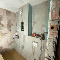 DOMINIX & ORIANIX & TRAIDING SPÓŁKA Z OGRANICZONĄ ODPOWIEDZIALNOŚCIĄ - Łazienka w trakcie wykończenia: marmurowe płytki na ścianach, widoczne instalacje hydrauliczne i narzędzia glazurnicze. Fragment ściany z cegły w surowym stanie.