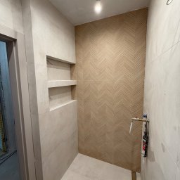 Wykończona łazienka z beżowymi płytkami ułożonymi w jodełkę na ścianie, wnęki w ścianie wyłożone płytkami, widoczna instalacja wodna i odpływ liniowy.