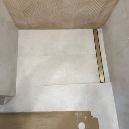 Wykończona łazienka z beżowymi płytkami na ścianach i podłodze, z odpływem liniowym w kolorze złotym. Na podłodze leżą kartony i wiadro glazurnicze.