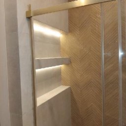 DOMINIX & ORIANIX & TRAIDING SPÓŁKA Z OGRANICZONĄ ODPOWIEDZIALNOŚCIĄ - Nowoczesna kabina prysznicowa z podświetlanymi półkami wnękowymi i dekoracyjną ścianą z płytek ułożonych we wzór jodełki. Złote wykończenia dodają elegancji.