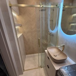 DOMINIX & ORIANIX & TRAIDING SPÓŁKA Z OGRANICZONĄ ODPOWIEDZIALNOŚCIĄ - Stylowa łazienka z kabiną prysznicową ze złotymi detalami, umywalką w odcieniach beżu i okrągłym lustrem z podświetleniem LED. Na ścianie wzór w jodełkę.