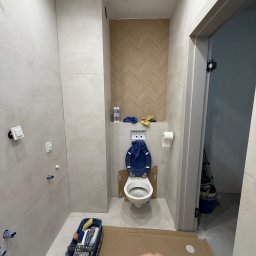 DOMINIX & ORIANIX & TRAIDING SPÓŁKA Z OGRANICZONĄ ODPOWIEDZIALNOŚCIĄ - Remont łazienki w trakcie: białe płytki, toaleta z niebieską deską, narzędzia w wiadrze, karton na podłodze. Widoczny fragment dekoracyjnej ściany z wzorem jodełki.