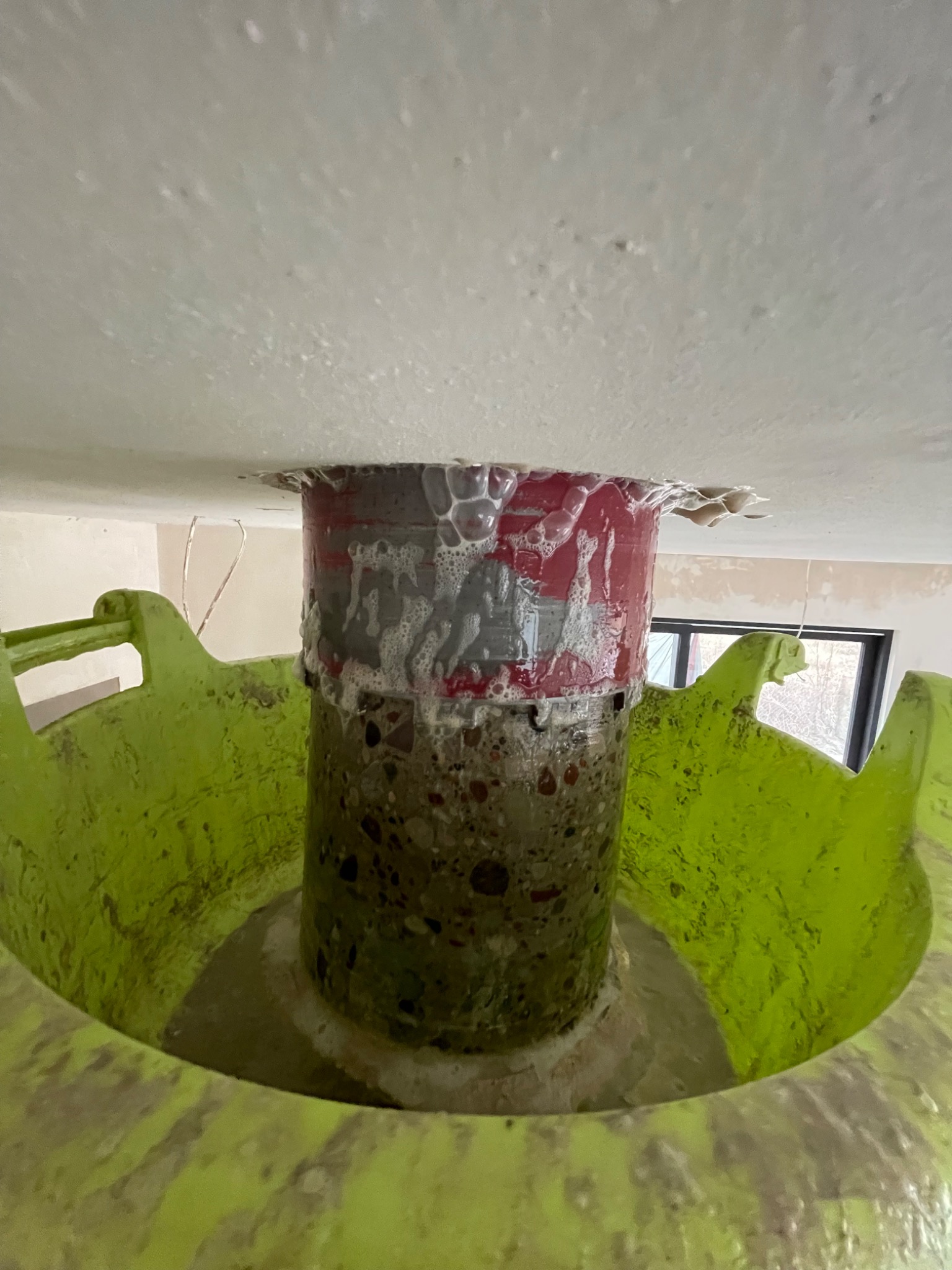 Próba szczelności połączenia rur w podłodze, widoczne ślady piany montażowej, zielona obudowa rury i surowy stan wykończenia pomieszczenia w tle.