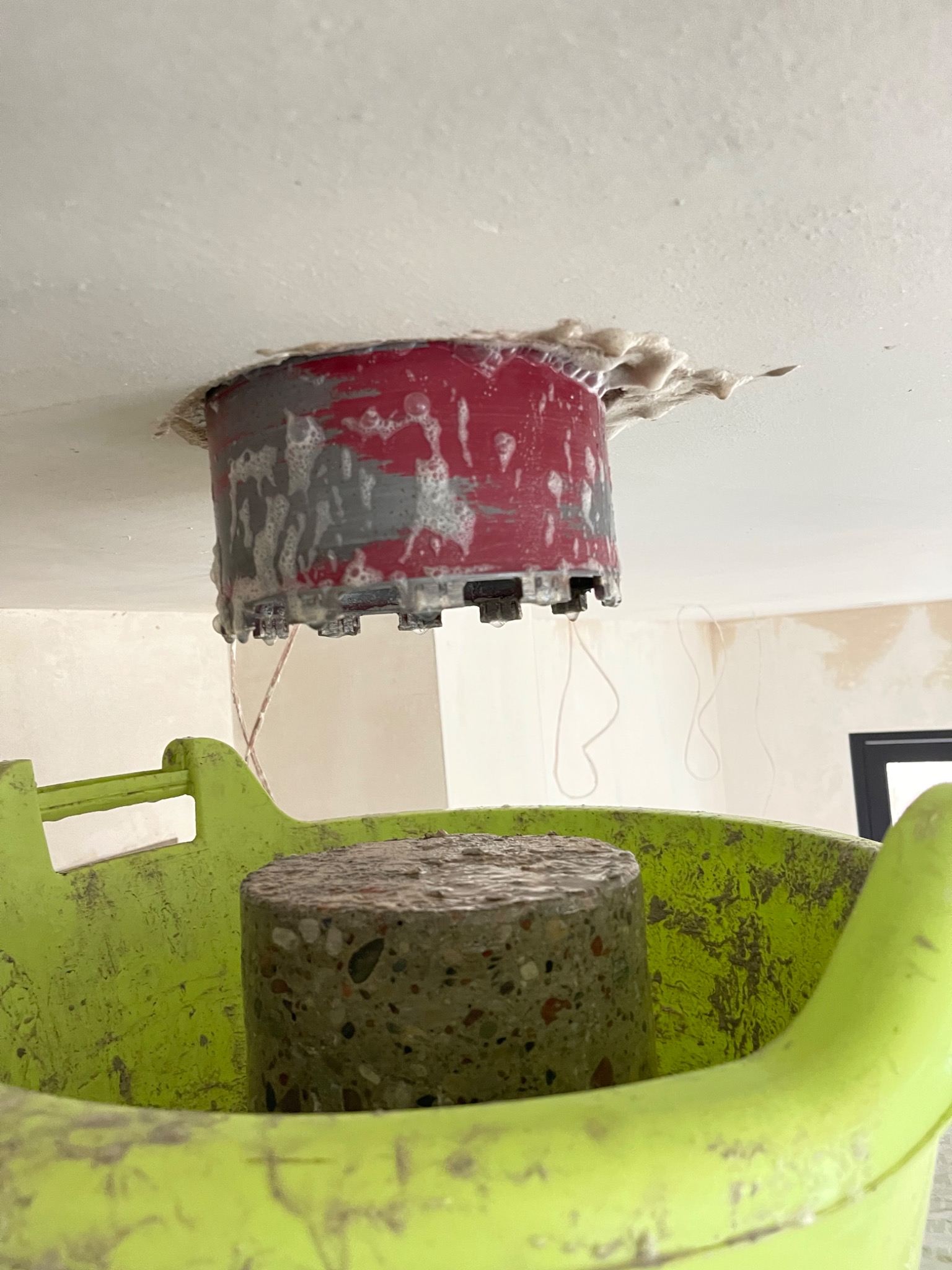 Wiertło koronowe z resztkami betonu po wierceniu otworu w suficie, umieszczone w zielonym wiadrze.