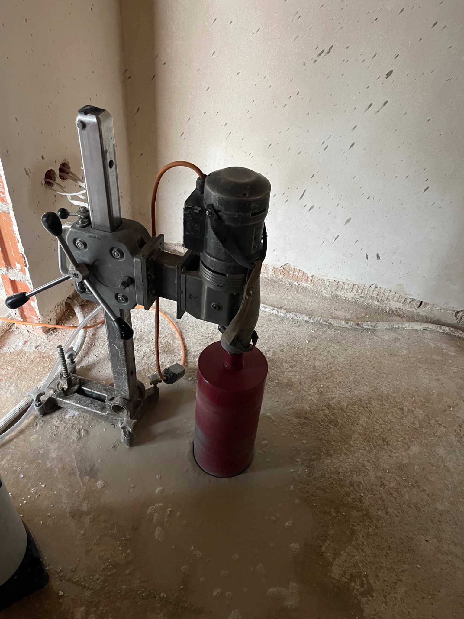 Wiertnica diamentowa z czerwoną koronką w trakcie pracy, widoczne ślady wiercenia w ścianie i woda na podłodze, surowe wykończenie wnętrza.