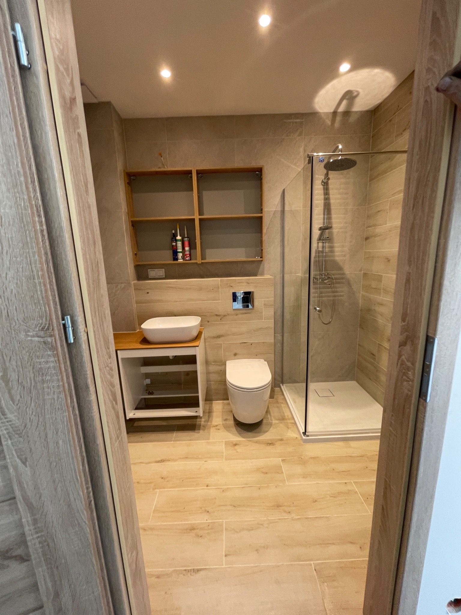 Jasna łazienka z kabiną prysznicową typu walk-in, umywalką nablatową na szafce i wiszącą toaletą. Na ścianie półki z kosmetykami i płytki imitujące drewno.