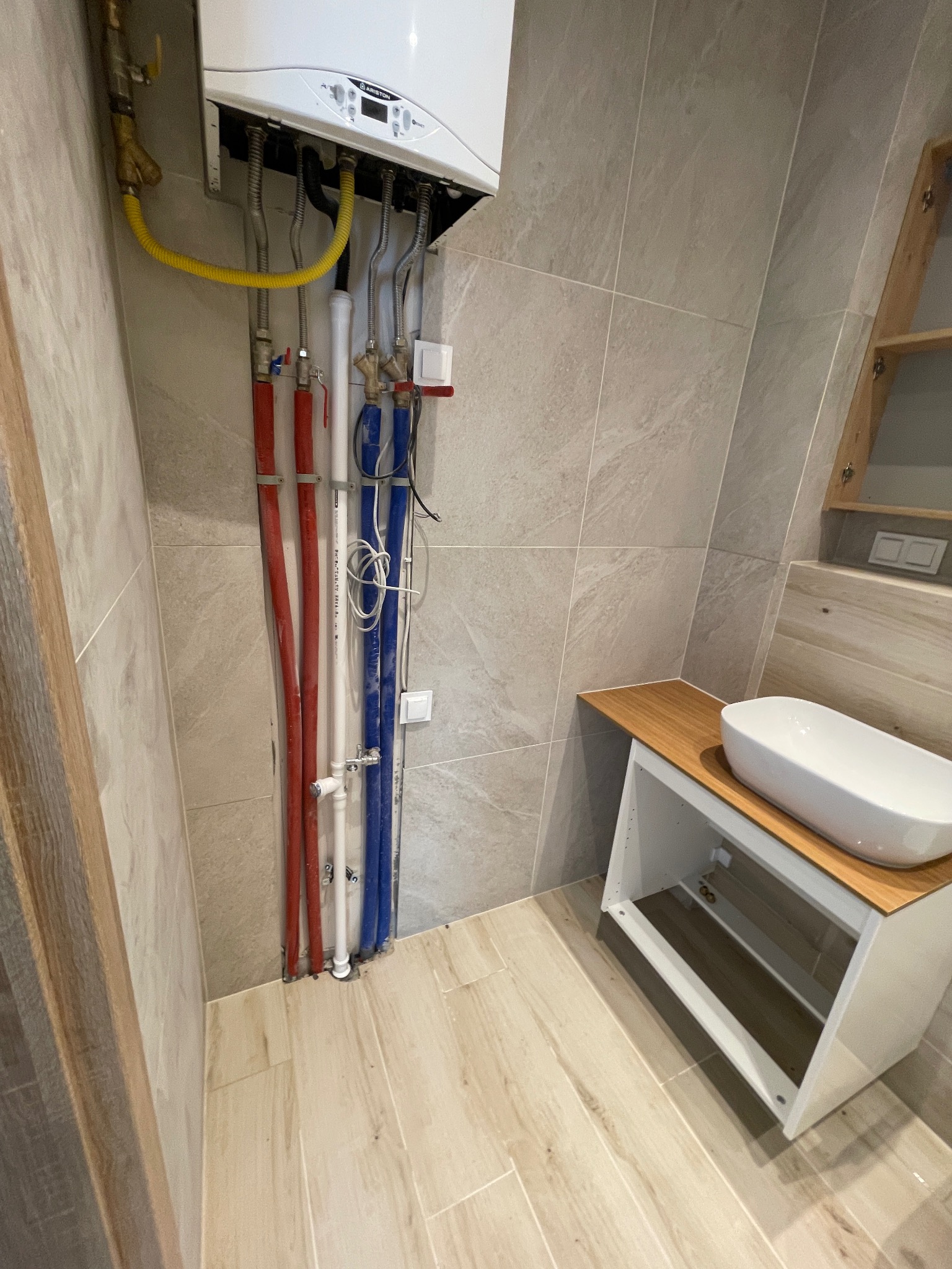Nowoczesna łazienka po remoncie: wiszący piec gazowy z instalacją hydrauliczną (czerwone i niebieskie rury), umywalka nablatowa na szafce z drewnianym blatem.