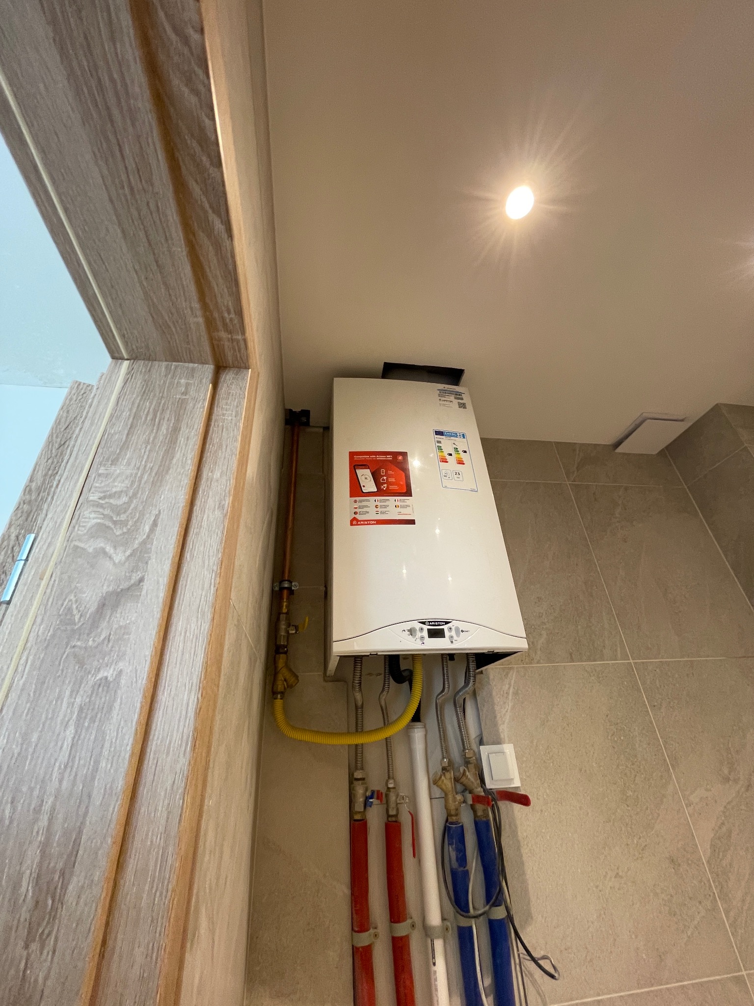 Biały, wiszący piec gazowy Ariston w rogu łazienki z beżowymi płytkami. Widoczne rury instalacyjne w kolorach czerwonym i niebieskim. Oświetlenie sufitowe.