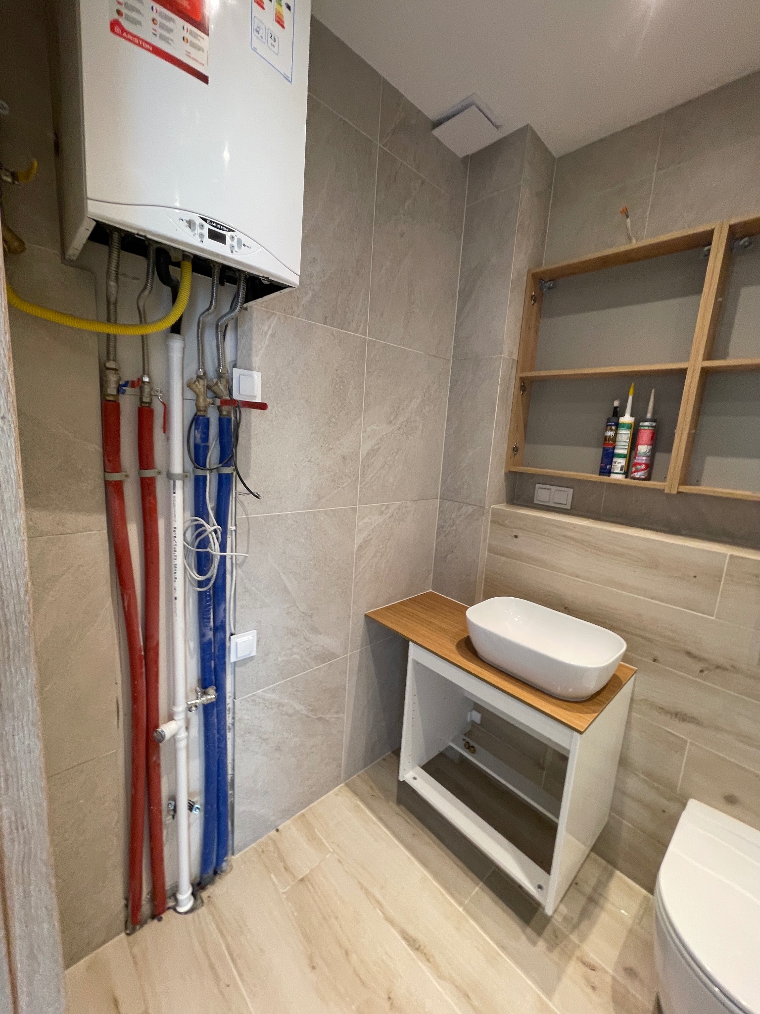 Nowoczesna łazienka w trakcie remontu: widoczny piec gazowy, instalacja hydrauliczna z rurami w kolorach czerwonym i niebieskim oraz umywalka na szafce z drewnianym blatem.
