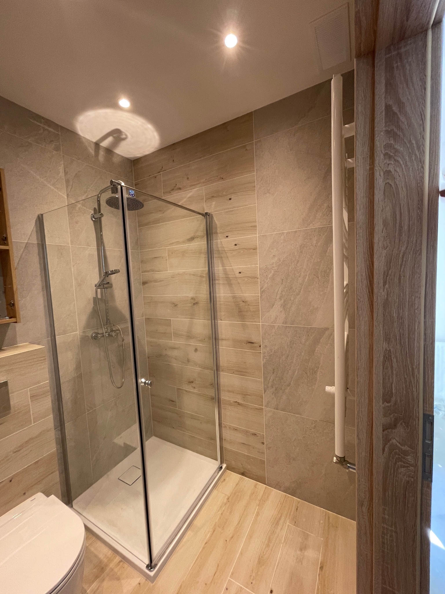 Nowoczesna kabina prysznicowa z deszczownicą, obok toaleta i grzejnik drabinkowy. Płytki imitujące drewno na ścianach i podłodze. Jasne, ciepłe oświetlenie.