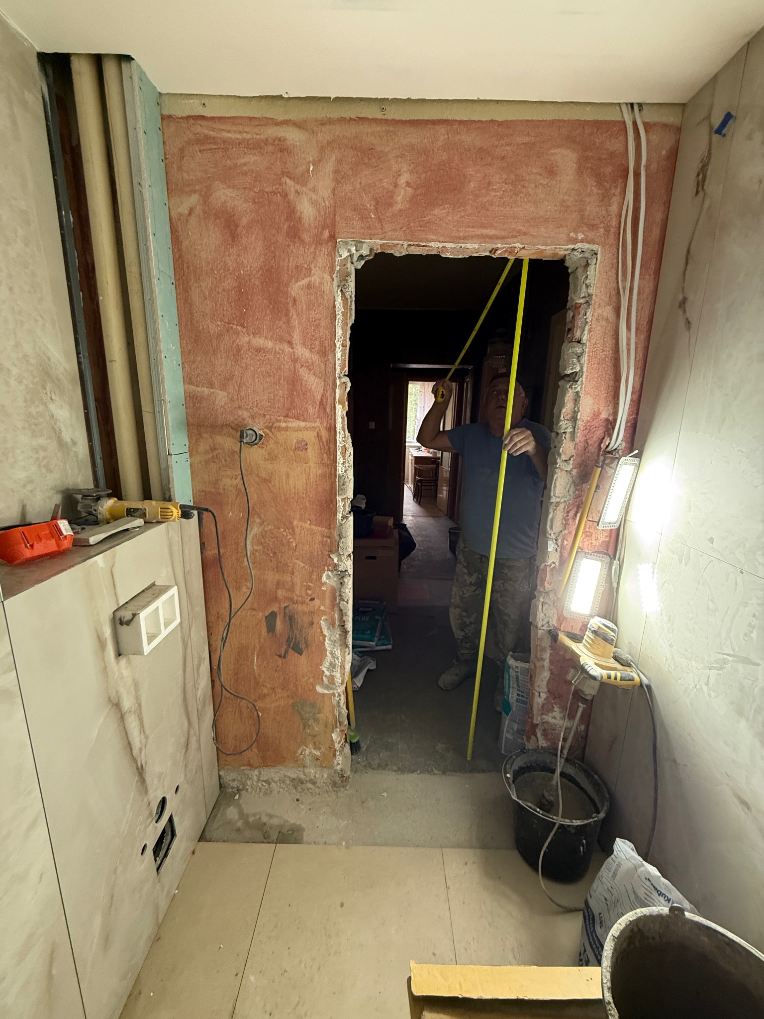 Remont łazienki w trakcie: mężczyzna mierzy otwór drzwiowy taśmą mierniczą. Widoczne narzędzia, kafelki na ścianach i podłodze, ściany w trakcie przygotowania.