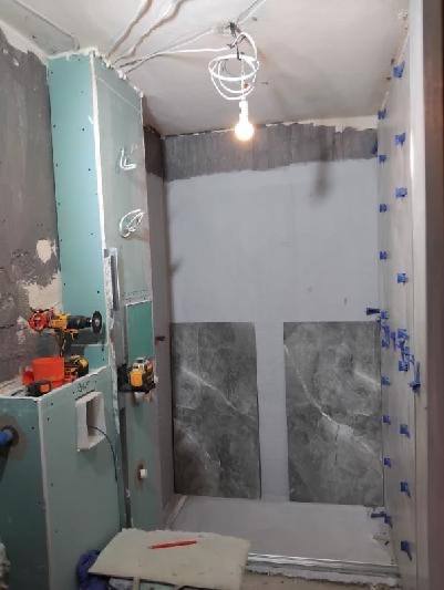 Łazienka w trakcie remontu: płyty gipsowe, narzędzia, częściowo ułożone płytki z niebieskimi krzyżykami dystansowymi. Widoczne przewody elektryczne.