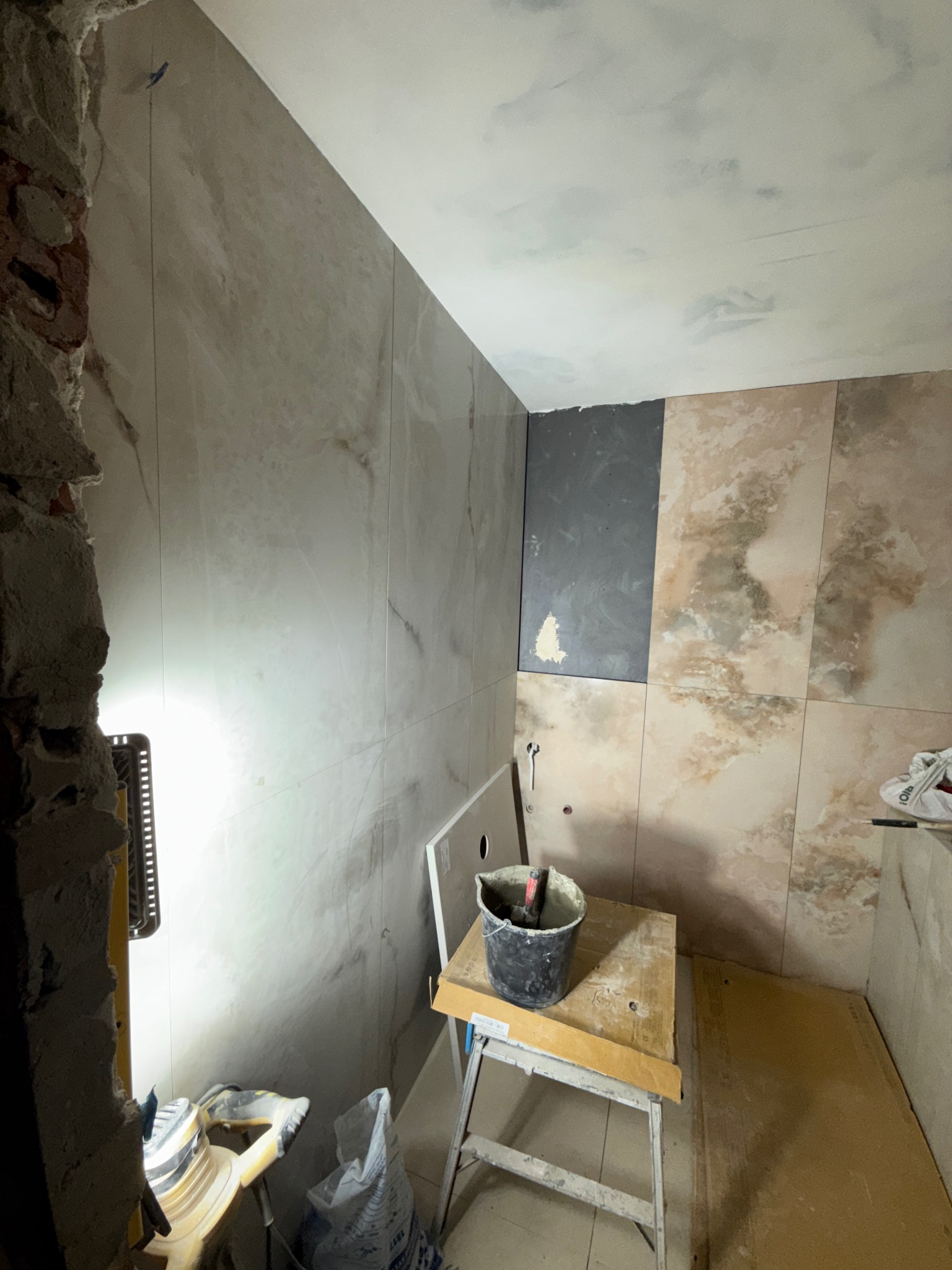 Wnętrze łazienki w trakcie remontu. Widoczne płytki imitujące marmur na ścianach, fragment ściany bez płytek, narzędzia i wiadro na podeście. Oświetlenie halogenowe.