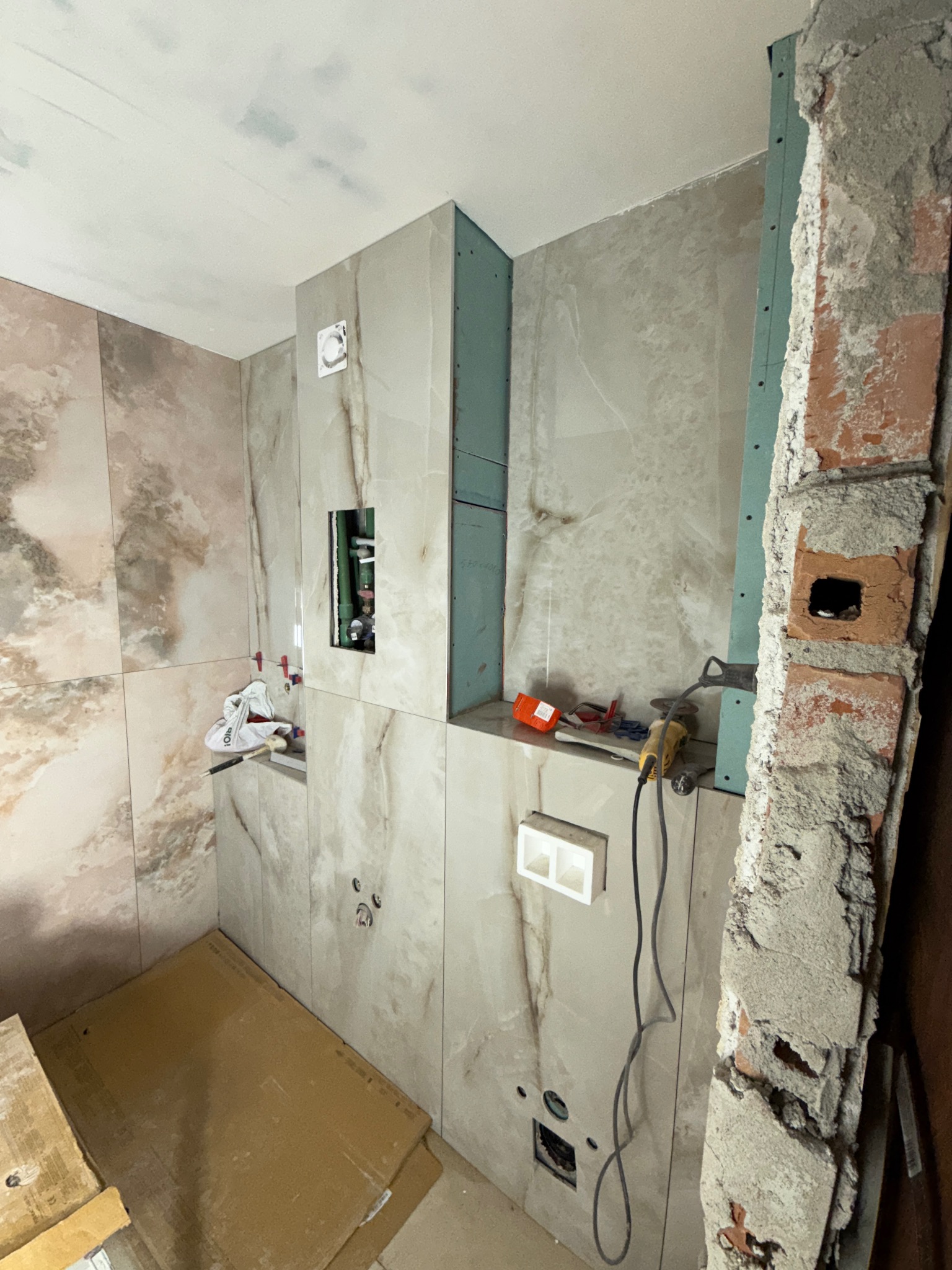 Łazienka w trakcie wykończenia: marmurowe płytki na ścianach, widoczne instalacje hydrauliczne i narzędzia glazurnicze. Fragment ściany z cegły w surowym stanie.