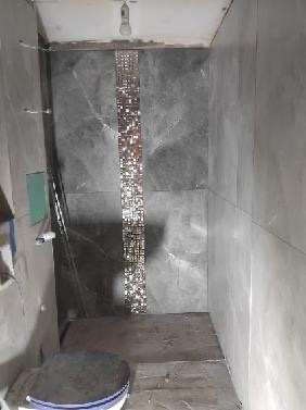 Wykończona ściana łazienki z szarymi płytkami i mozaiką dekoracyjną. Widoczne niedokończone ściany i podłoga. Ujęcie z dołu, surowy stan wykończenia.