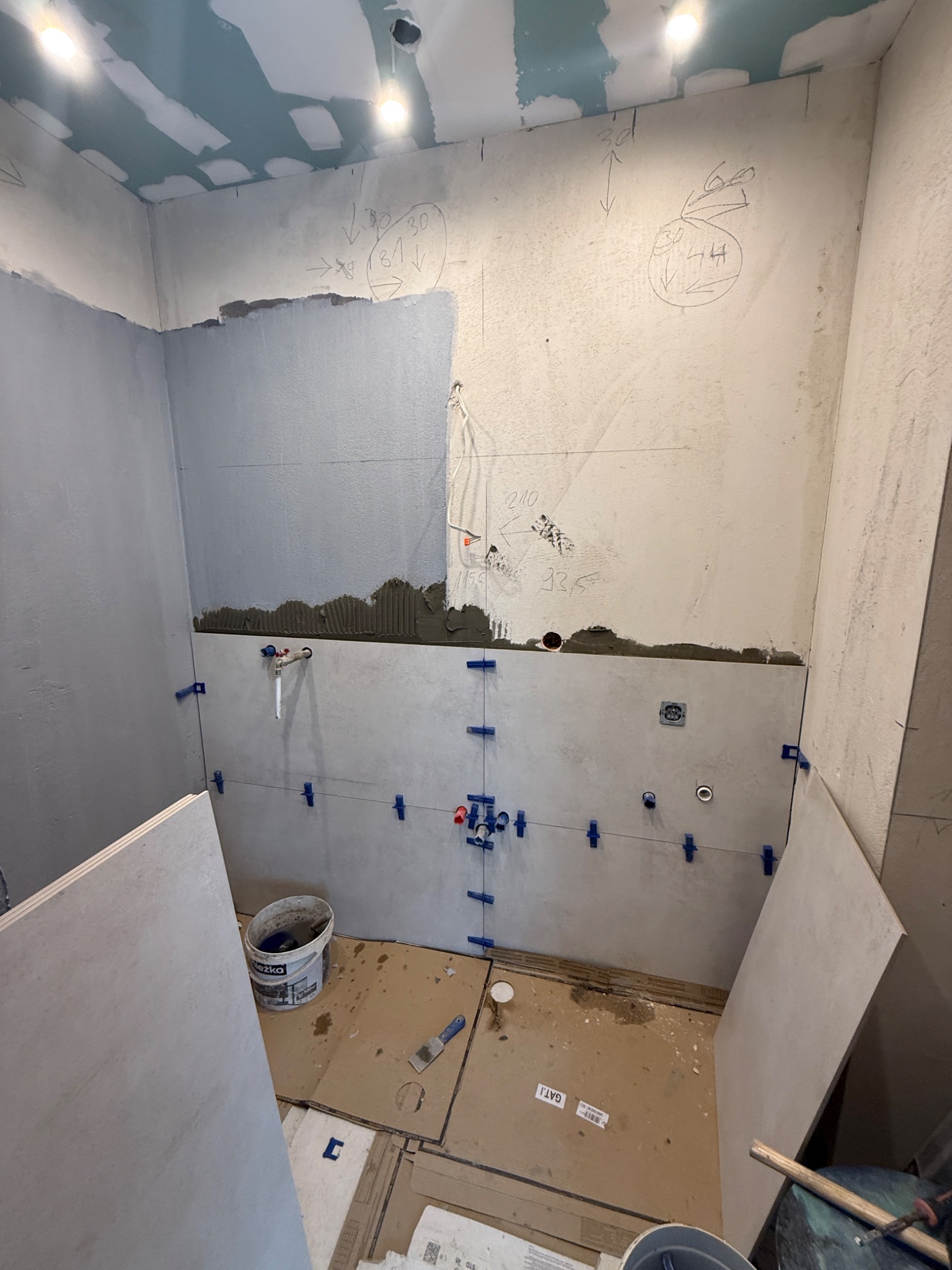 Wnętrze łazienki w trakcie remontu. Widoczne płytki na ścianach, poziomica, wiadra i narzędzia glazurnicze. Na ścianach zaznaczone wymiary i punkty instalacji.