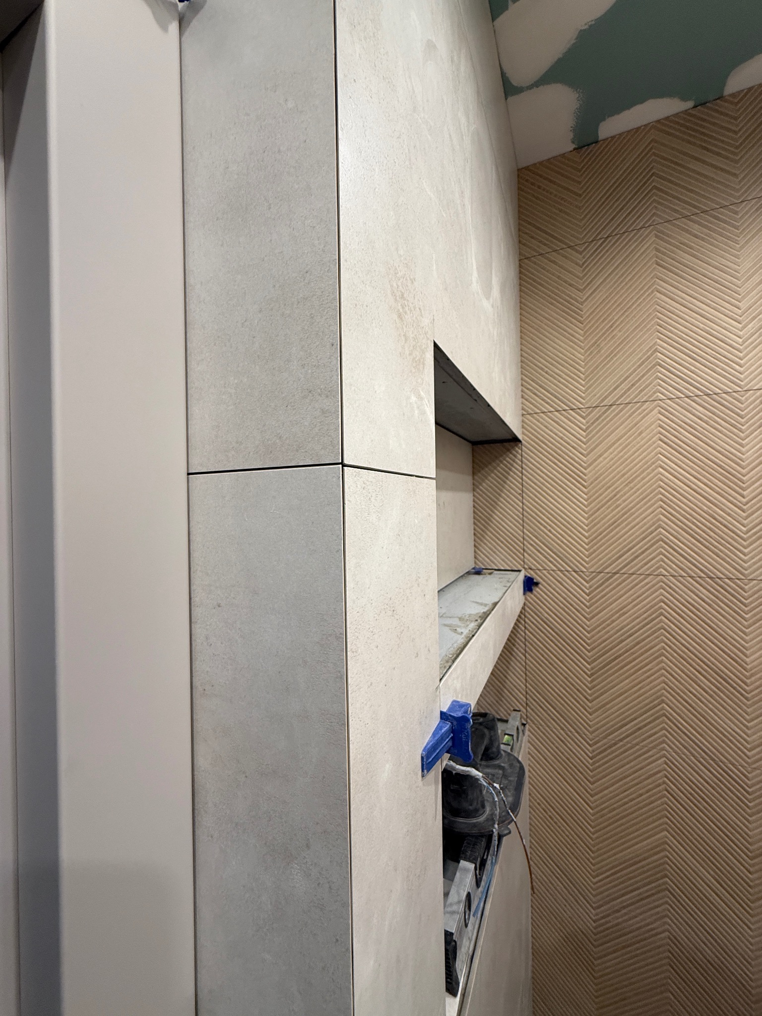 Ściana łazienki w trakcie wykańczania z beżowymi płytkami ułożonymi we wzór jodełki i wnęką wyłożoną szarymi płytkami. Widoczne narzędzia glazurnicze.