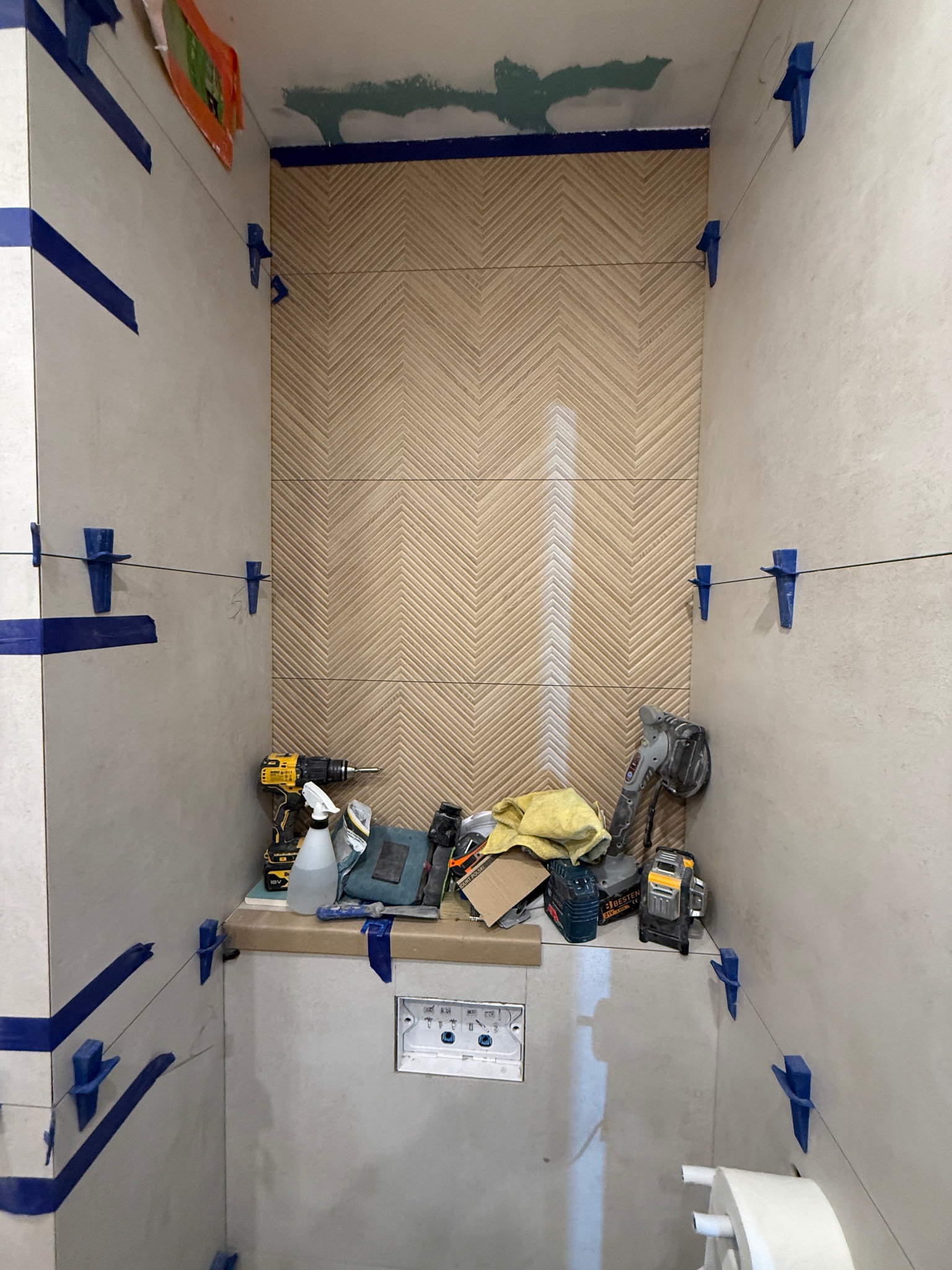 Układanie płytek w łazience z użyciem poziomicy laserowej i klipsów. Na półce widoczne narzędzia: wiertarka, szlifierka kątowa i akcesoria glazurnicze.