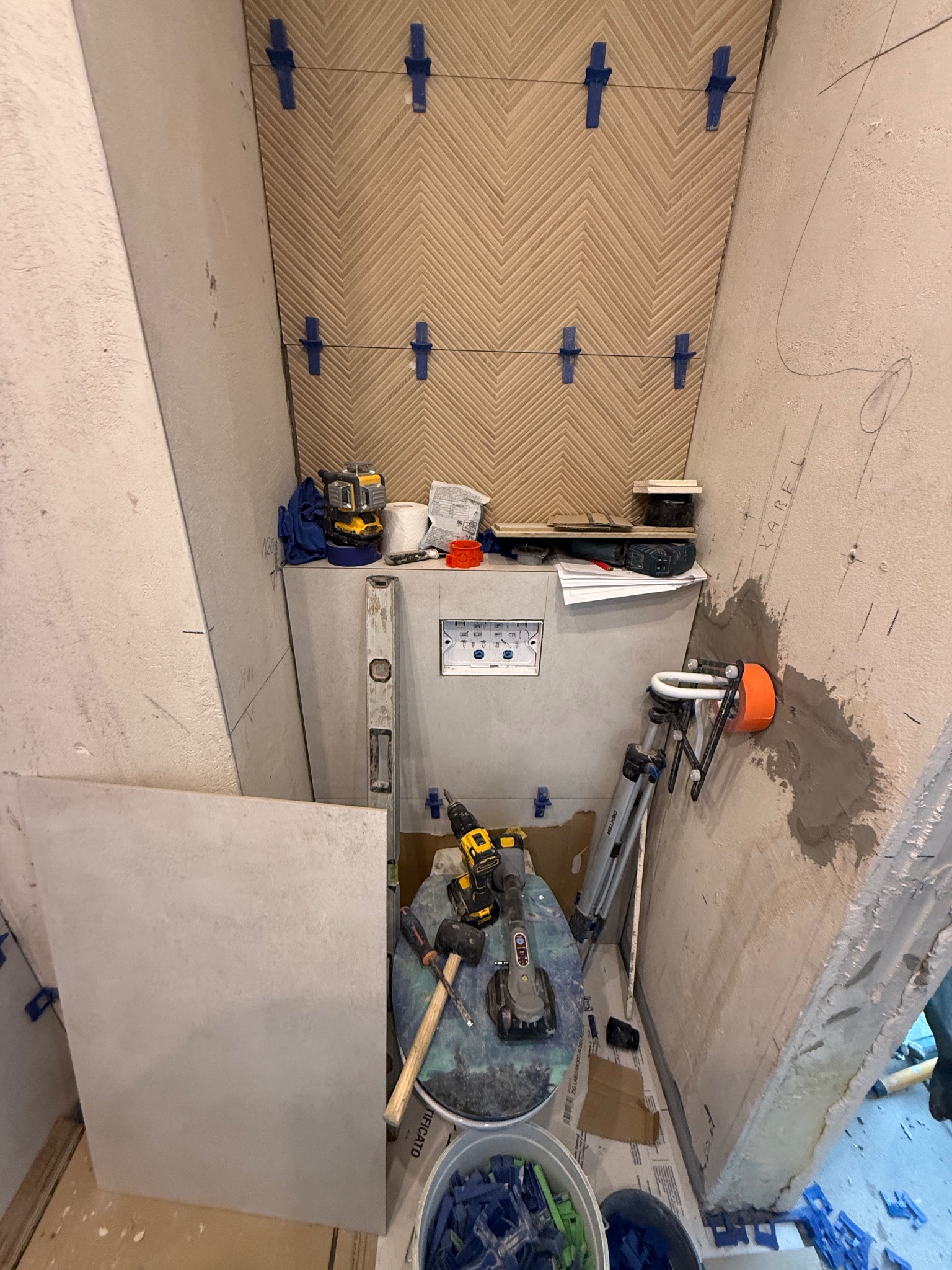 Wąska łazienka w trakcie wykończenia: ułożona częściowo wzorzysta glazura na ścianie, narzędzia glazurnicze, poziomica,  w tle instalacja podtynkowa WC.