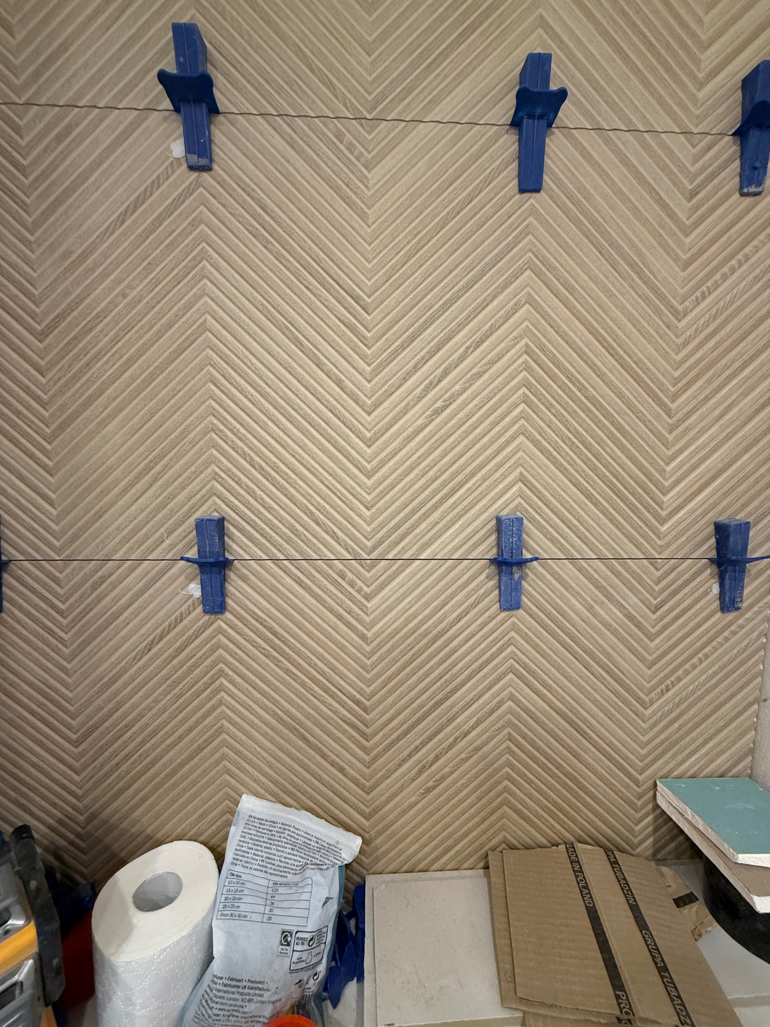 Ułożona glazura w jodełkę, z niebieskimi klipsami poziomującymi. Widoczne materiały budowlane i narzędzia. Detal z realizacji wykończenia wnętrz.