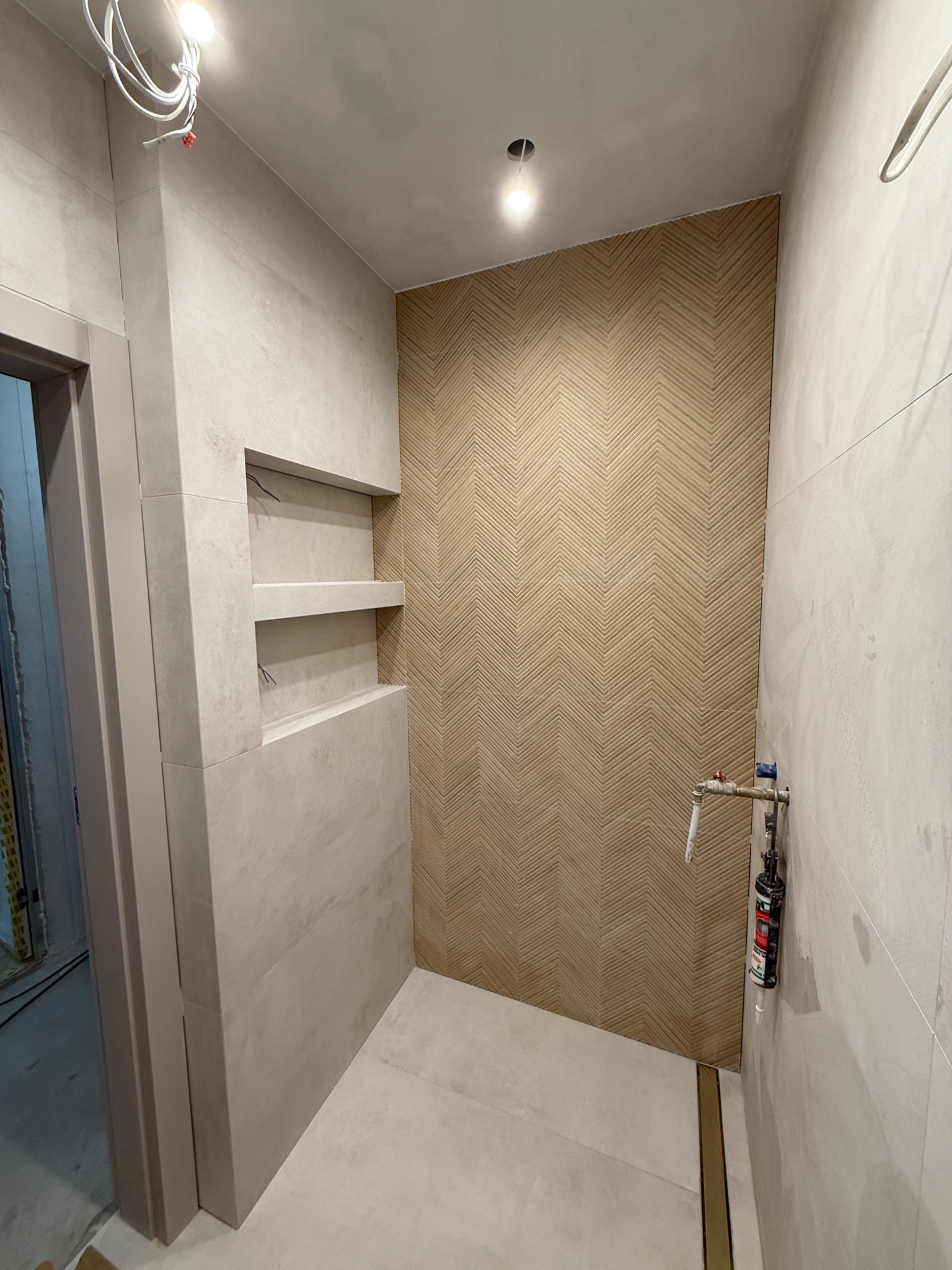 Wykończona łazienka z beżowymi płytkami ułożonymi w jodełkę na ścianie, wnęki w ścianie wyłożone płytkami, widoczna instalacja wodna i odpływ liniowy.