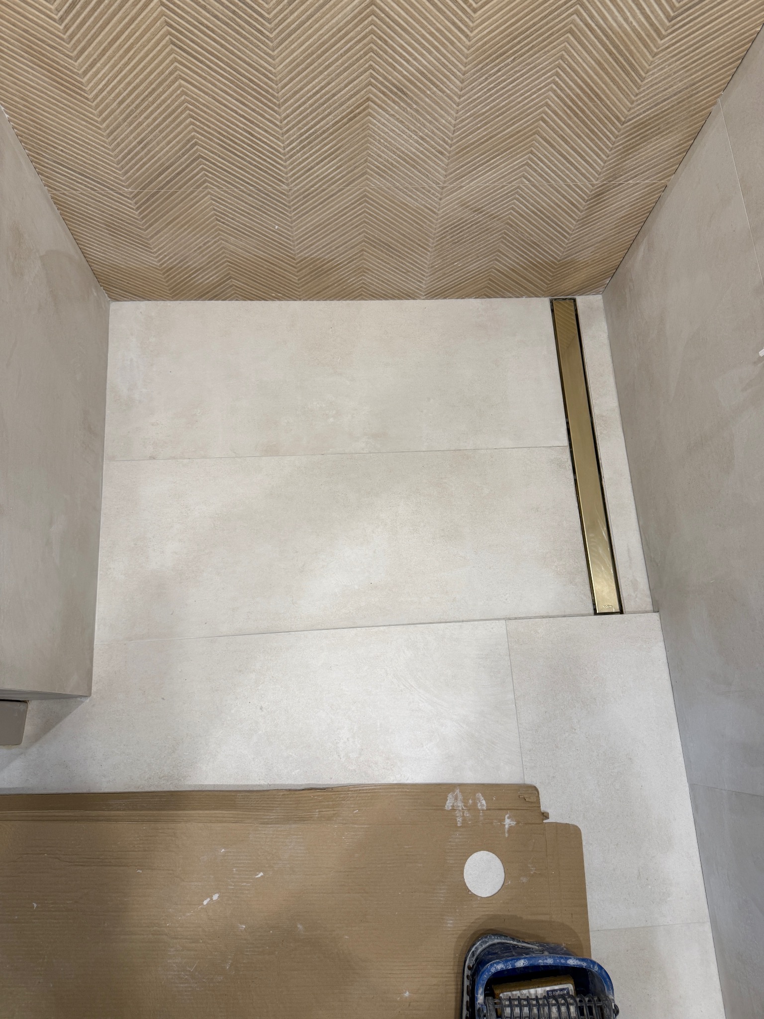 Wykończona łazienka z beżowymi płytkami na ścianach i podłodze, z odpływem liniowym w kolorze złotym. Na podłodze leżą kartony i wiadro glazurnicze.