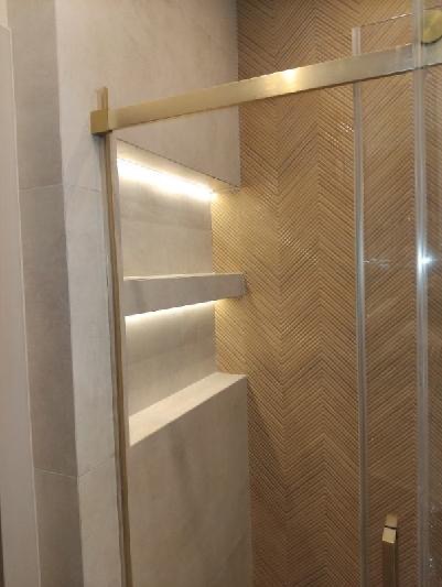 Nowoczesna kabina prysznicowa z podświetlanymi półkami wnękowymi i dekoracyjną ścianą z płytek ułożonych we wzór jodełki. Złote wykończenia dodają elegancji.