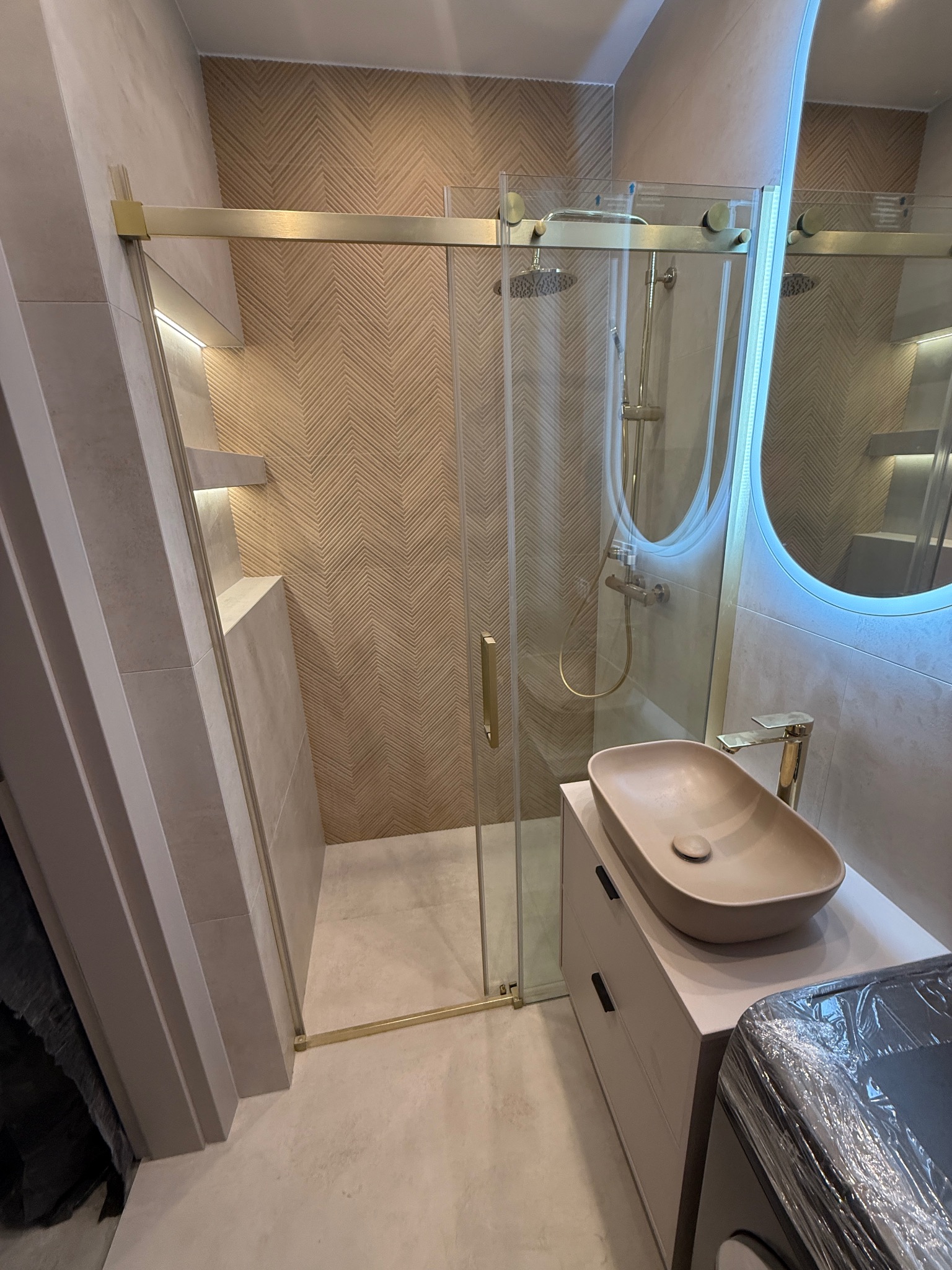 Stylowa łazienka z kabiną prysznicową ze złotymi detalami, umywalką w odcieniach beżu i okrągłym lustrem z podświetleniem LED. Na ścianie wzór w jodełkę.
