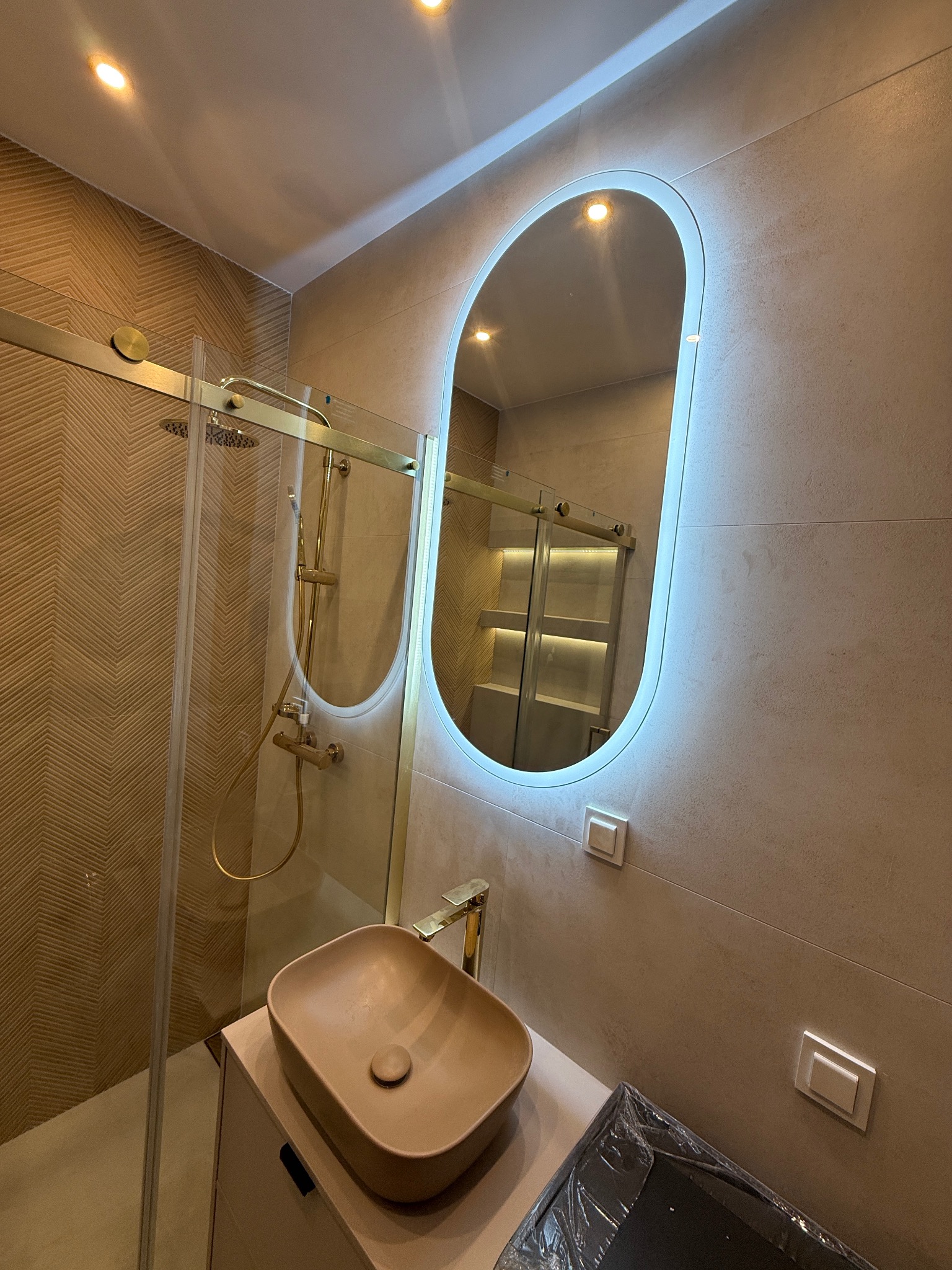 Stylowa łazienka w Warszawie: beżowe płytki, kabina prysznicowa ze złotymi elementami i owalne lustro z podświetleniem LED nad umywalką.