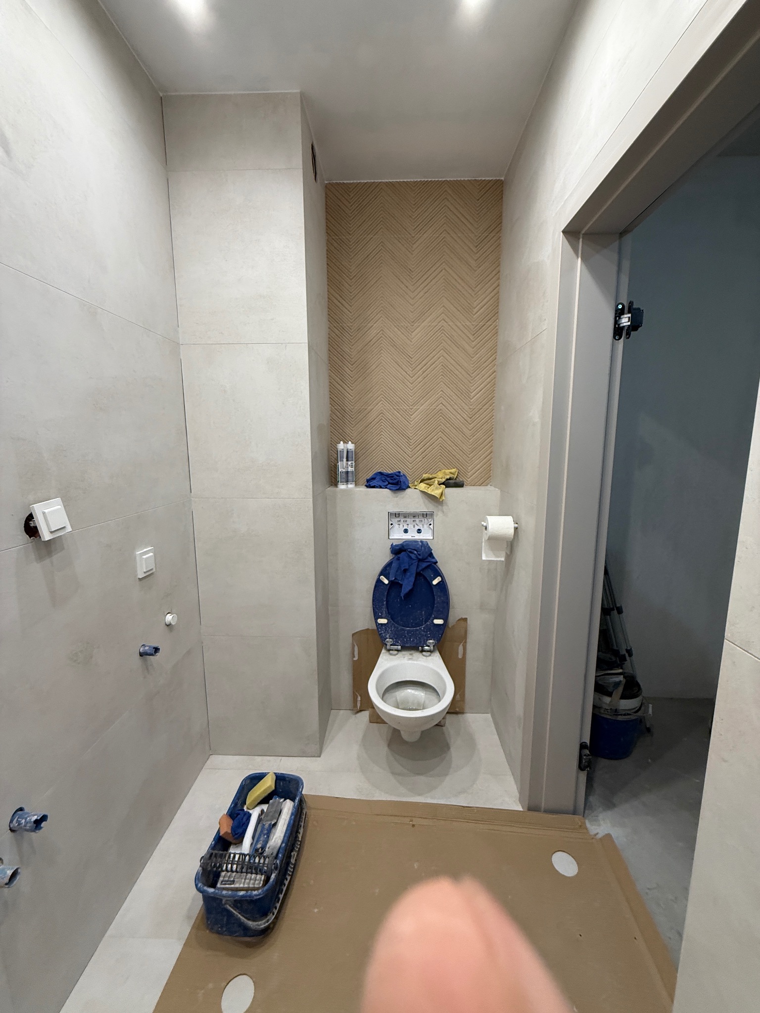 Remont łazienki w trakcie: białe płytki, toaleta z niebieską deską, narzędzia w wiadrze, karton na podłodze. Widoczny fragment dekoracyjnej ściany z wzorem jodełki.