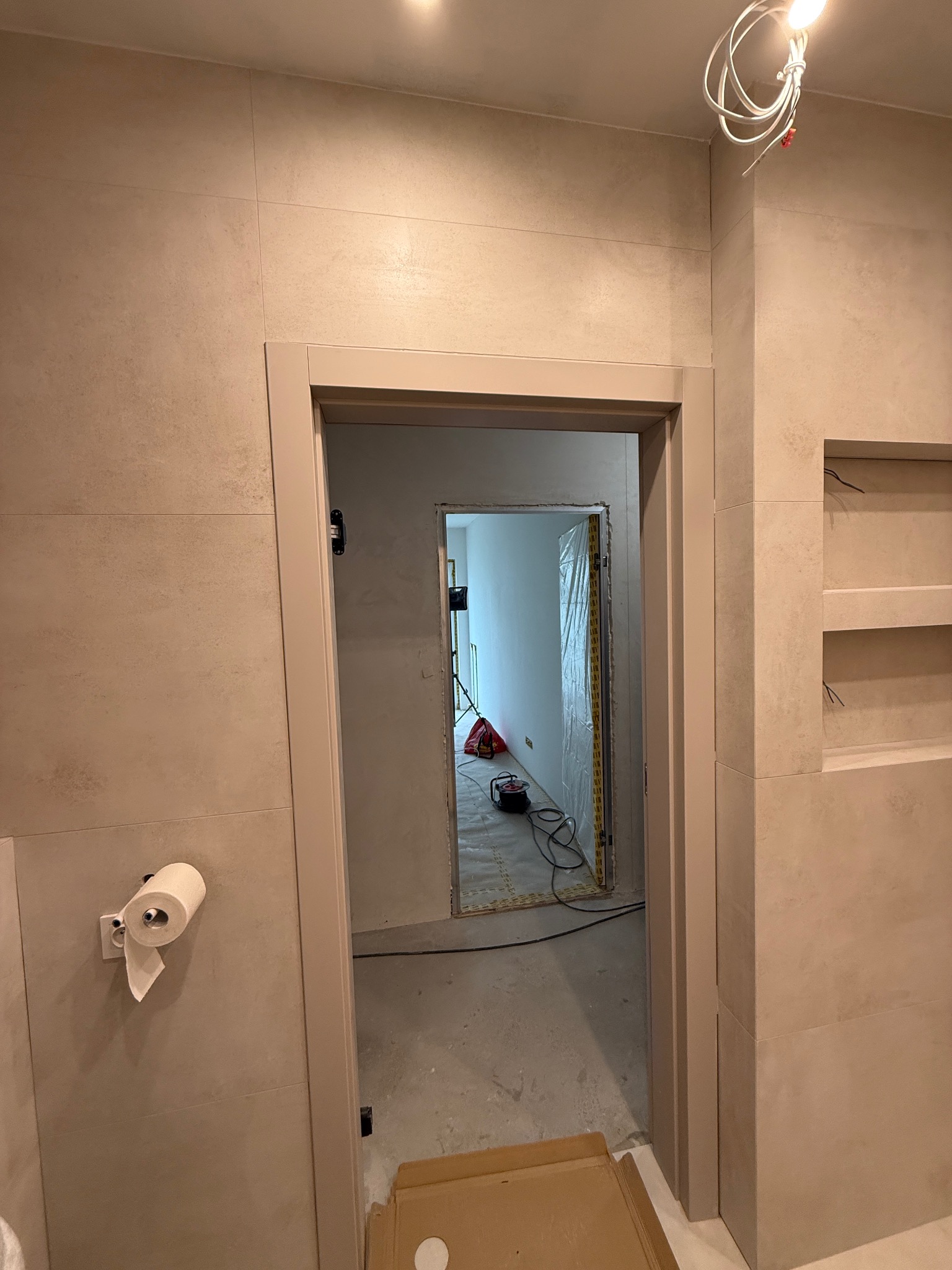 Beżowa łazienka w trakcie wykończenia: widoczne płytki, wnęka z półkami, zamontowana rama drzwiowa i rolka papieru toaletowego. W tle narzędzia i folia ochronna.