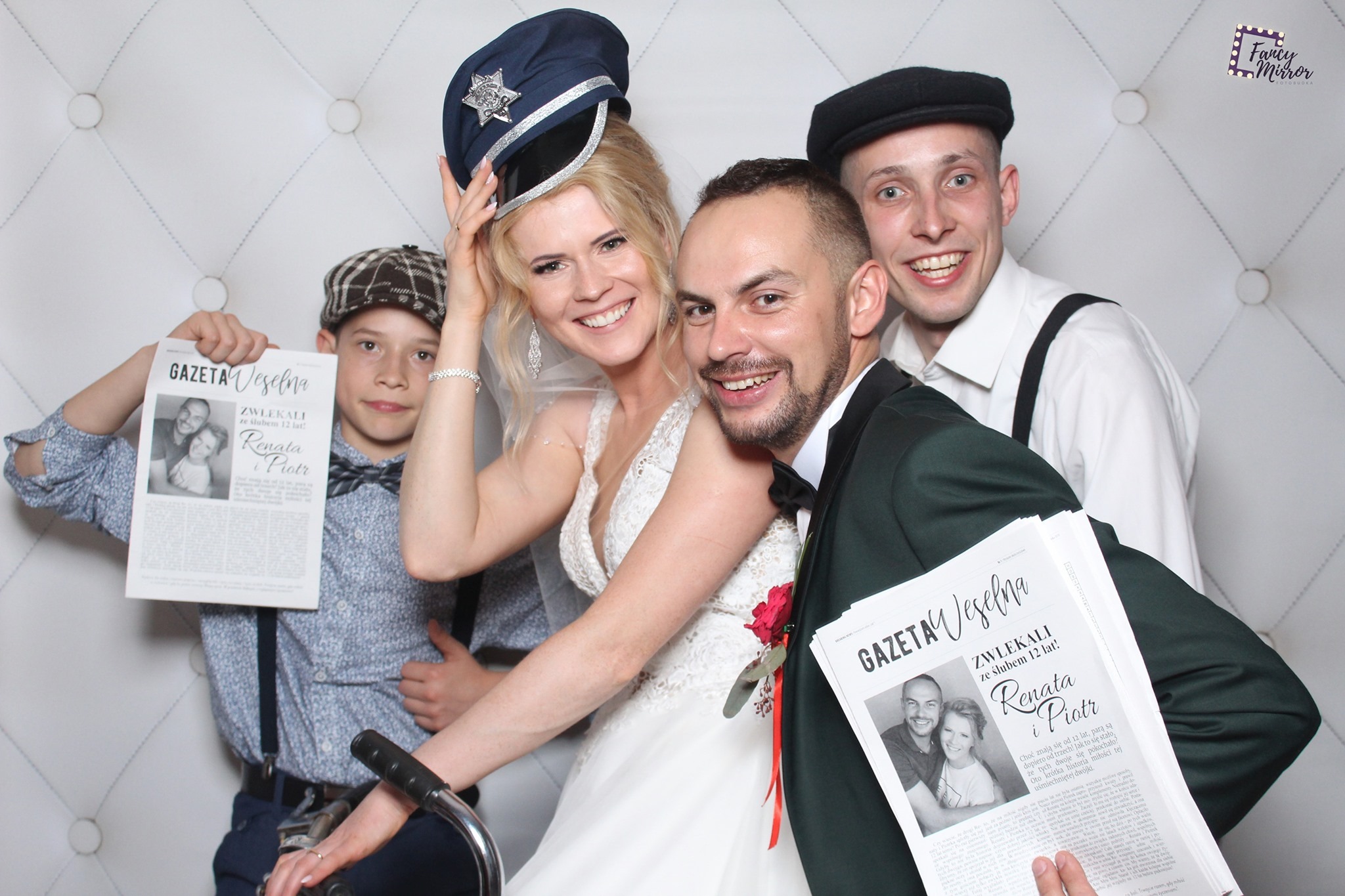 Zdjęcie z fotobudki: uśmiechnięta para młoda z gośćmi trzymająca rekwizyty - gazety weselne z ich zdjęciem, panna młoda w policyjnej czapce, tło pikowane.