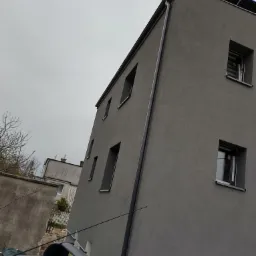 Szara elewacja budynku z oknami i rynnami, widok z dołu z perspektywy dachu samochodu, pochmurne niebo w tle.