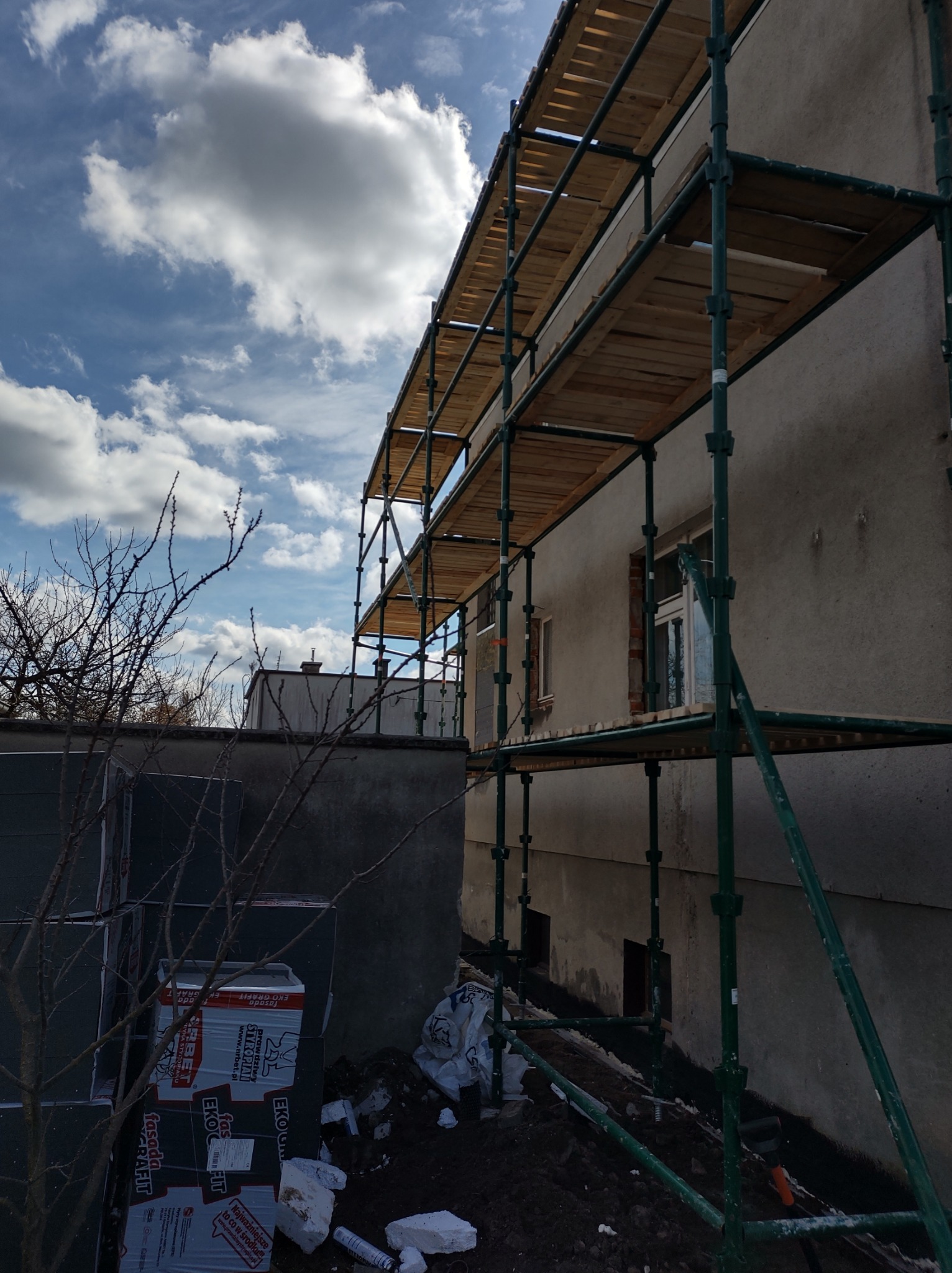 Budynek w trakcie prac elewacyjnych, widoczne rusztowania, materiały izolacyjne i fragmenty odsłoniętej cegły wokół okna.