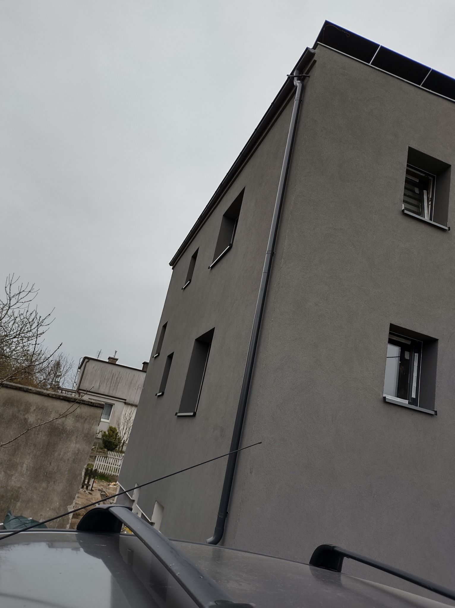 Szara elewacja budynku z oknami i rynnami, widok z dołu z perspektywy dachu samochodu, pochmurne niebo w tle.