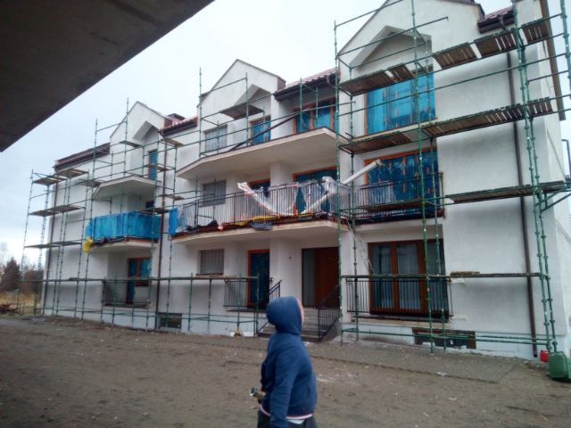 Trzypiętrowy budynek mieszkalny w trakcie prac elewacyjnych, zabezpieczony rusztowaniami i folią ochronną na oknach, widziany z perspektywy osoby w niebieskiej kurtce.