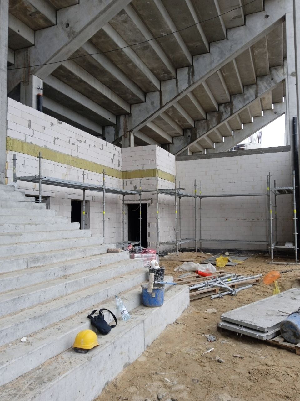 Budowa trybun stadionu: surowa konstrukcja betonowa, białe bloczki z ociepleniem, rusztowania, narzędzia i kask na schodach.
