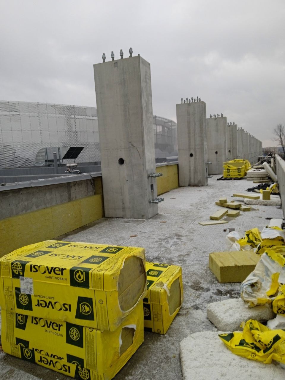 Montaż ocieplenia na elewacji budynku przemysłowego, widoczne betonowe słupy konstrukcyjne, żółte płyty izolacyjne Isover w opakowaniach i fragmenty ułożonego ocieplenia.