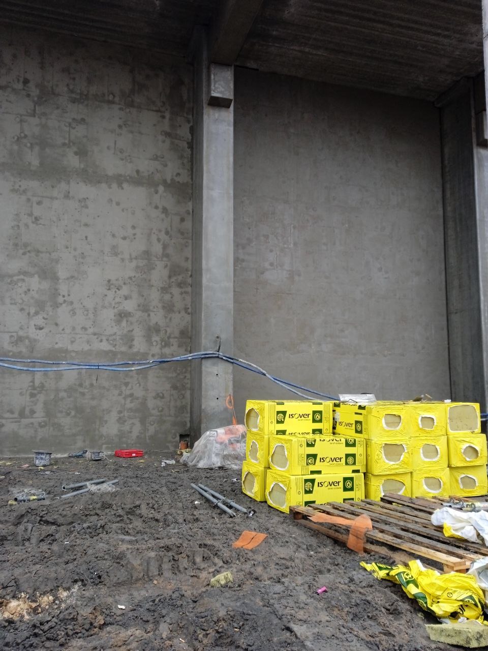 Surowa ściana budynku w trakcie przygotowań do ocieplenia, widoczne żółte paczki wełny mineralnej Isover na zabłoconym podłożu i niebieskie kable biegnące wzdłuż ściany.