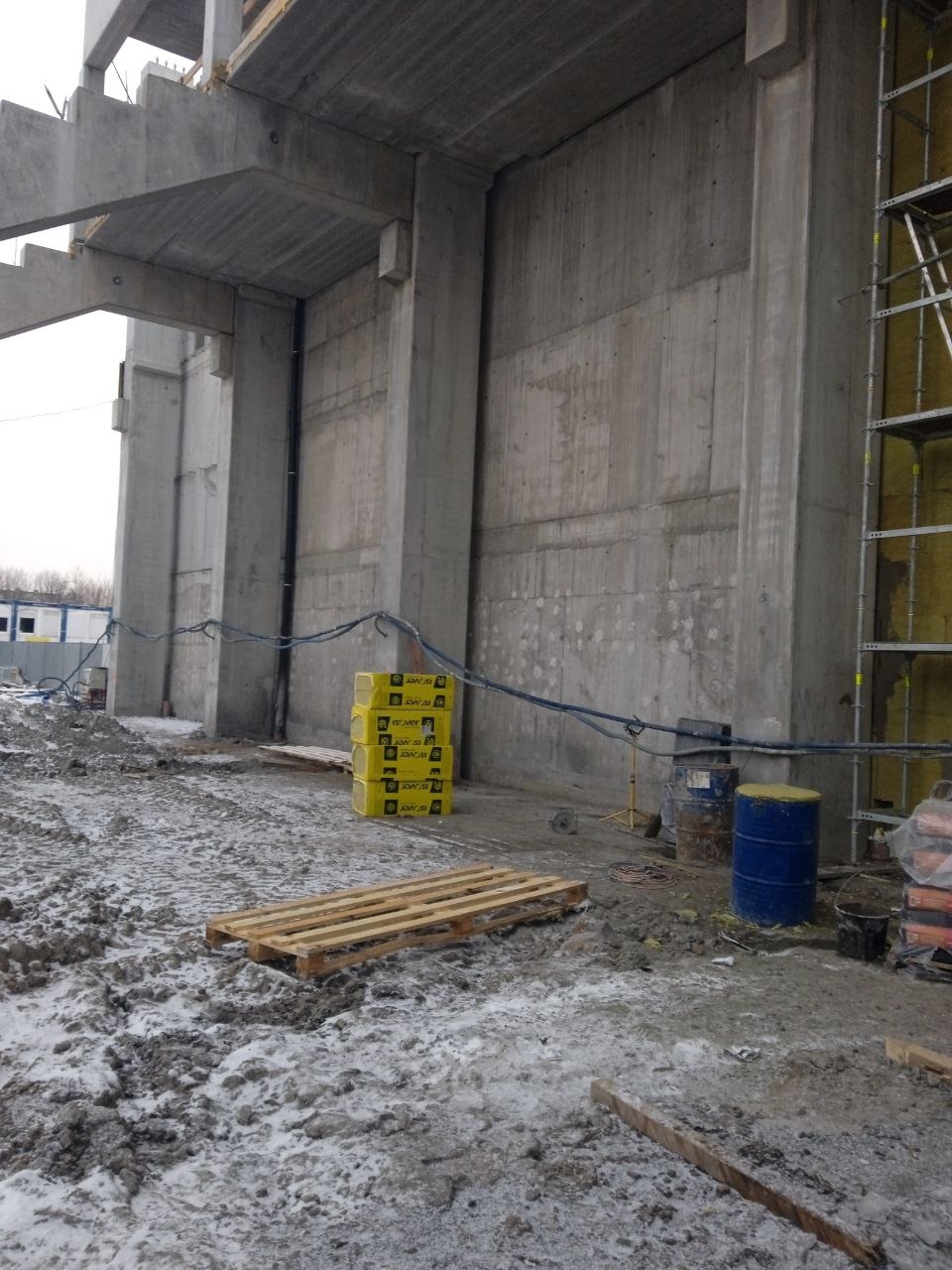 Surowa betonowa konstrukcja budynku w trakcie ocieplania, widoczne rusztowania, materiały izolacyjne w żółtych opakowaniach, paleta i beczki na zabłoconym placu budowy z resztkami śniegu.