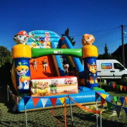 Kolorowy, dmuchany zamek z motywem Psiego Patrolu, dzieci zjeżdżają ze zjeżdżalni, na tle błękitne niebo i samochód dostawczy z napisem DMUCHAŃCE.