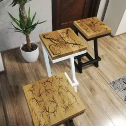 Trzy drewniane taborety z unikalnym wzorem przypominającym drzewo, wykonanym techniką Lichtenberga, na metalowych nogach w kolorze białym i czarnym, ustawione na drewnianej podłodze, obok doniczki...