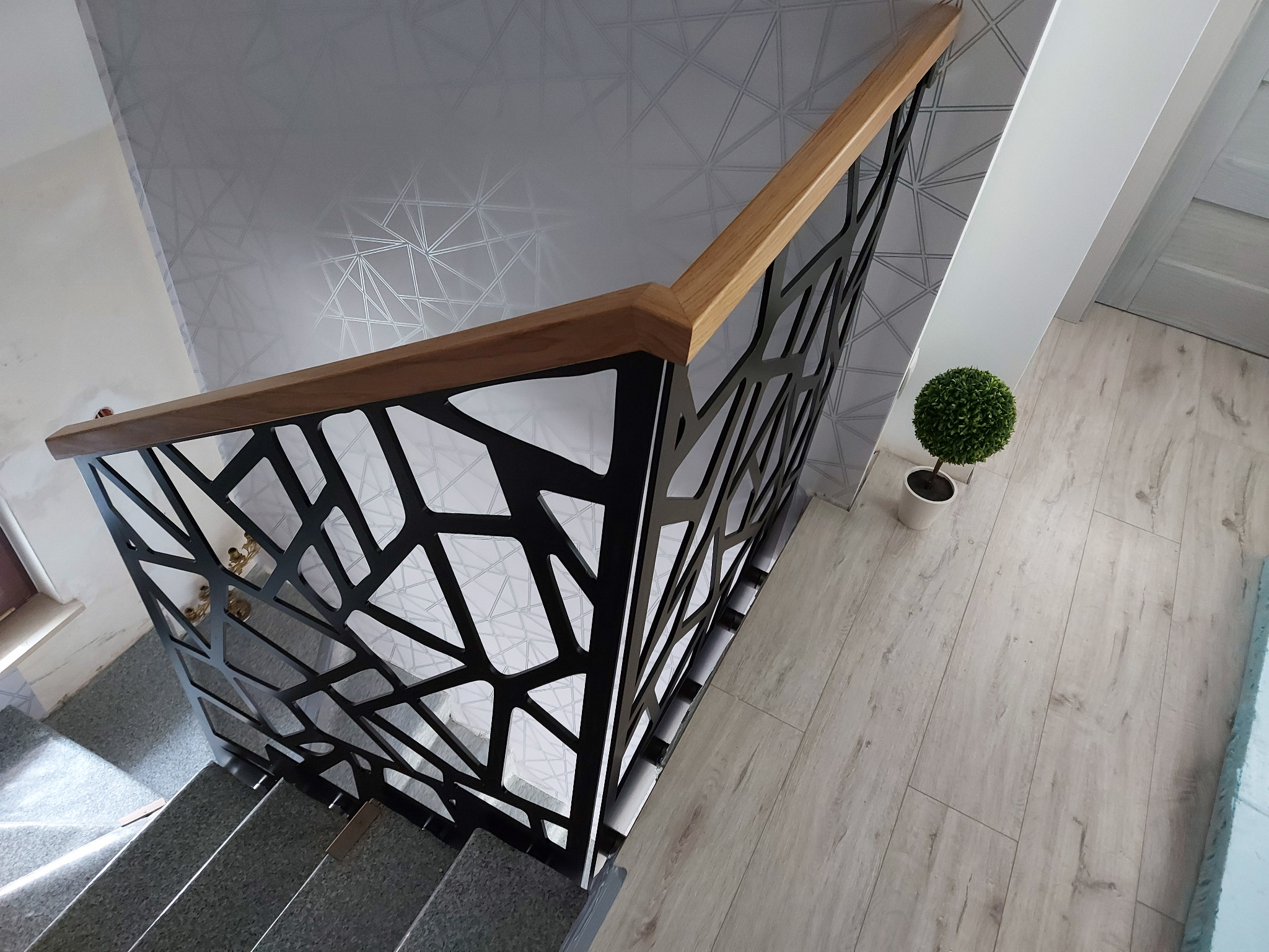 Nowoczesna balustrada schodowa z czarnym, geometrycznym wypełnieniem i drewnianym pochwytem, widziana z góry, z dekoracyjną tapetą w tle i małą rośliną doniczkową na podłodze.