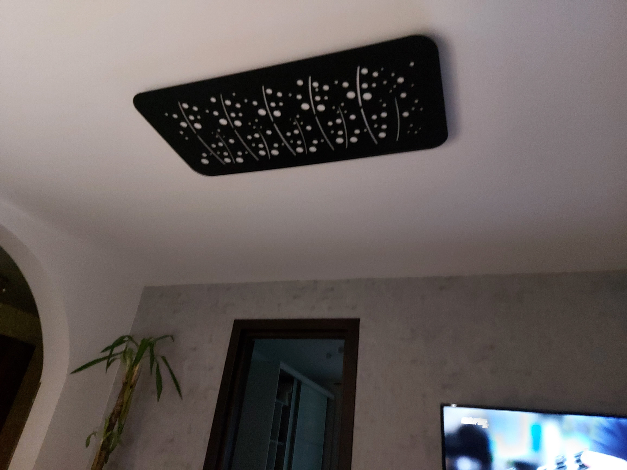 Czarna, prostokątna lampa sufitowa z wycinanym wzorem kropek i linii na białym suficie, widok z dołu, fragment ściany z rośliną i telewizorem w tle.