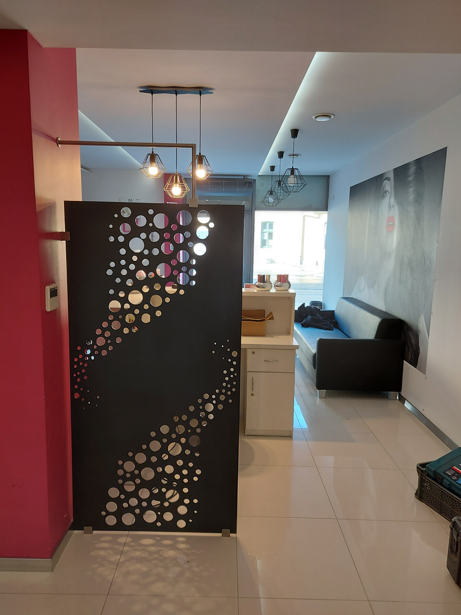 Czarny panel dekoracyjny z wyciętymi okręgami w salonie kosmetycznym, widoczne oświetlenie sufitowe, sofa i portret kobiety na ścianie.